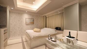 A modern hotel room with a bed, a glass coffee table with a bottle and glasses, a painting on the wall, and a ceiling light.