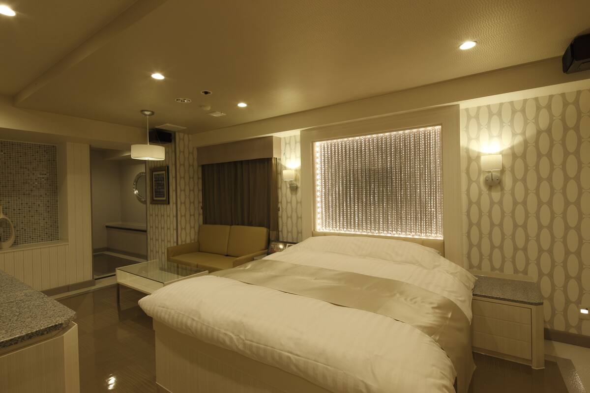 Un dormitorio moderno con una cama grande, una mesa de centro de vidrio, una zona de estar con sofá y un baño con ducha e inodoro.