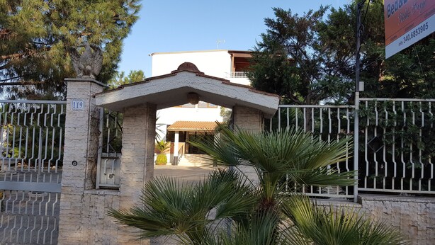 A gated entrance with a stone archway, a metal gate, and a building with a tiled roof in the background. - Villa Erry (Trani)