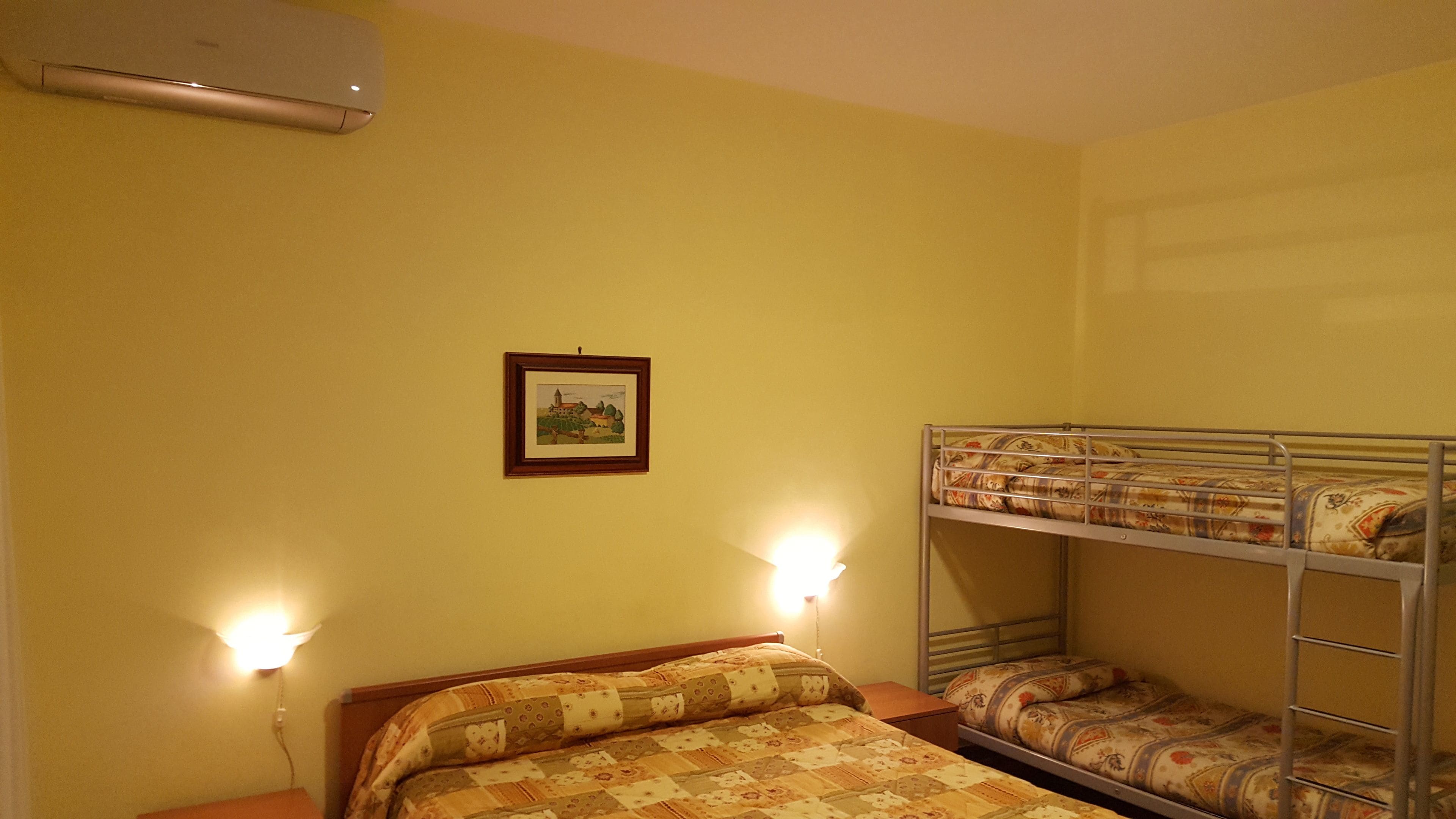 A dormitory room with bunk beds, a single bed, a framed picture on the wall, and two wall-mounted reading lights.