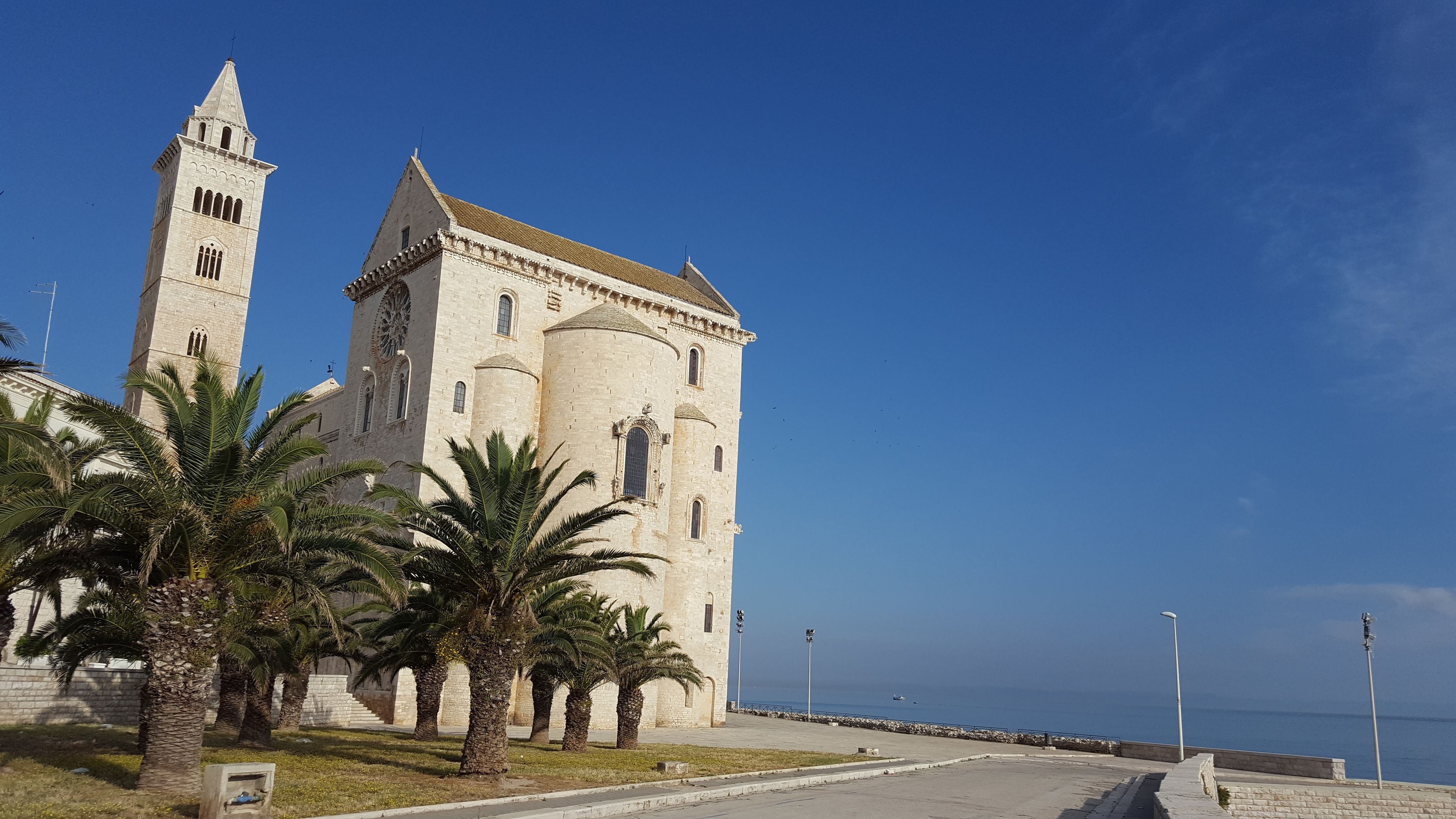 Un edificio histórico con una torre destacada y un amplio ventanal de arco, ubicado junto a una carretera costera con palmeras.
