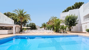 Una piscina con sillones blancos, palmeras y un edificio blanco con zona de bar.