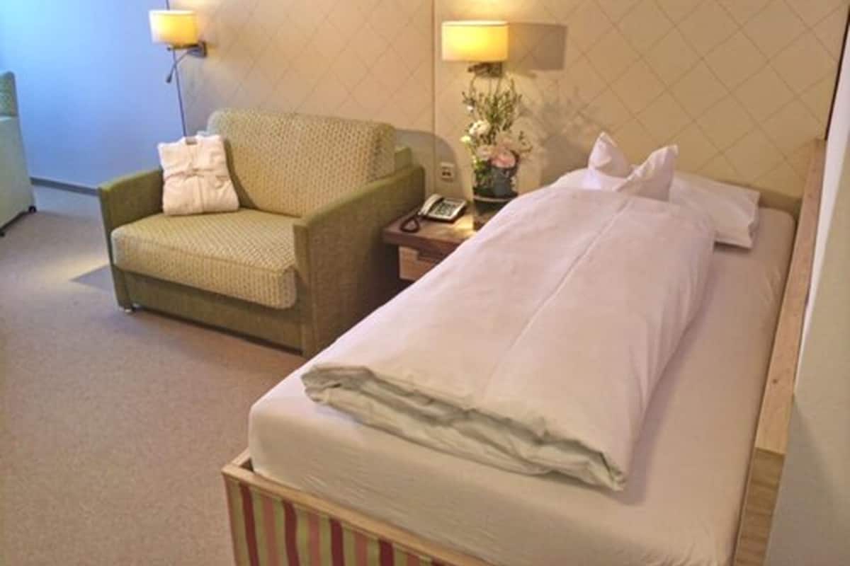 Quarto de hotel com uma cama de solteiro, uma cadeira, mesa de cabeceira com abajur e telefone.