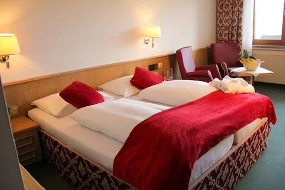 Quarto de hotel com uma cama de casal, duas almofadas, um cobertor vermelho, uma mesinha de cabeceira, uma luminária, uma cadeira e uma pequena mesa com uma cesta de frutas.