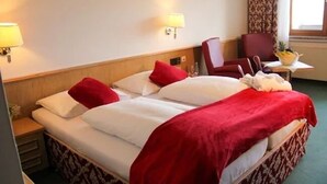 Ein Hotelzimmer mit Doppelbett, zwei Kissen, einer roten Tagesdecke, einem Nachttisch, einer Lampe, einem Sessel und einem kleinen Tisch mit Obstschale.