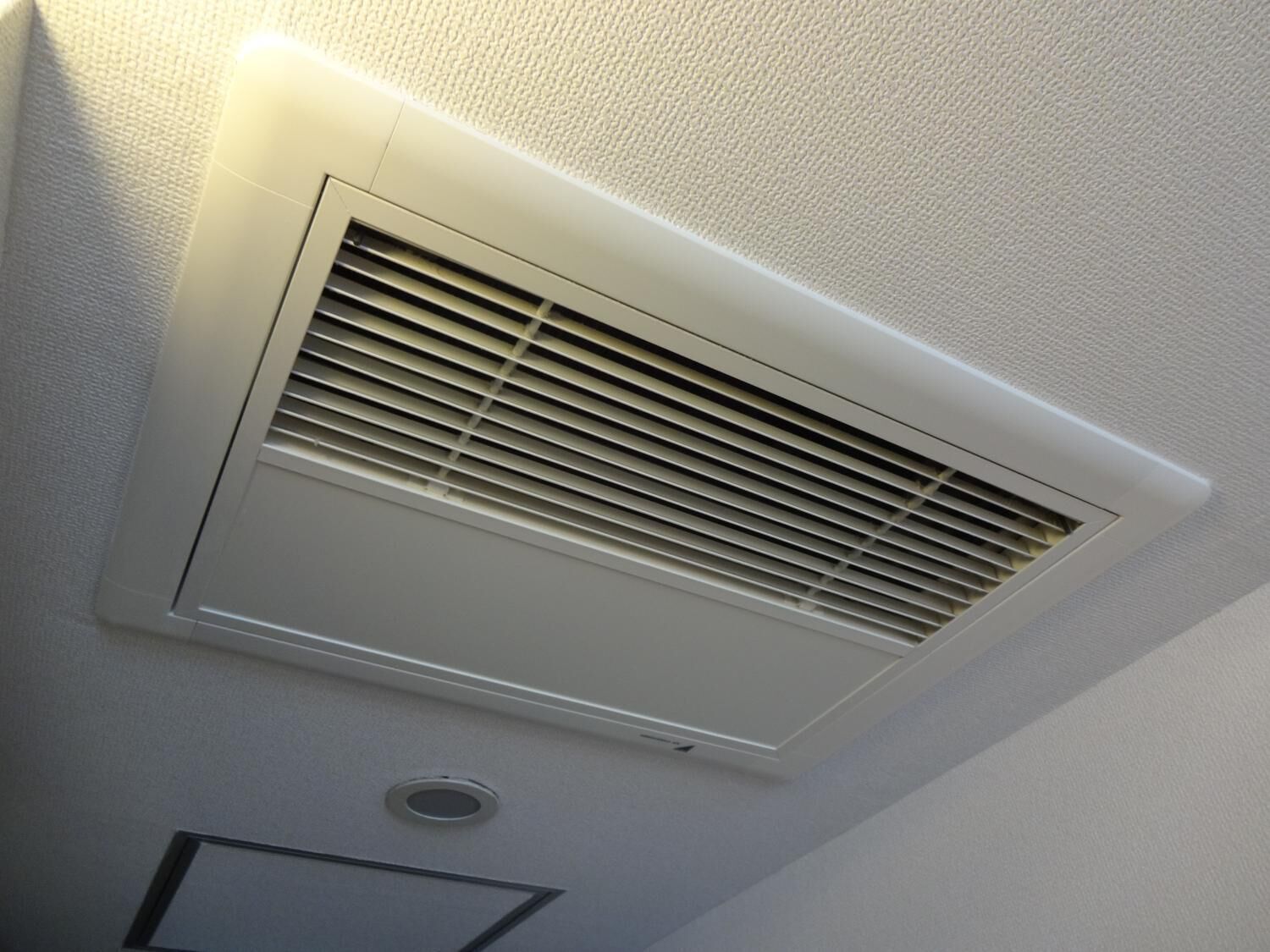 Et hvidt ventilationsgitter med vandrette lameller, et rundt ventilationsdæksel nedenunder, samt en indbygget lysarmatur.