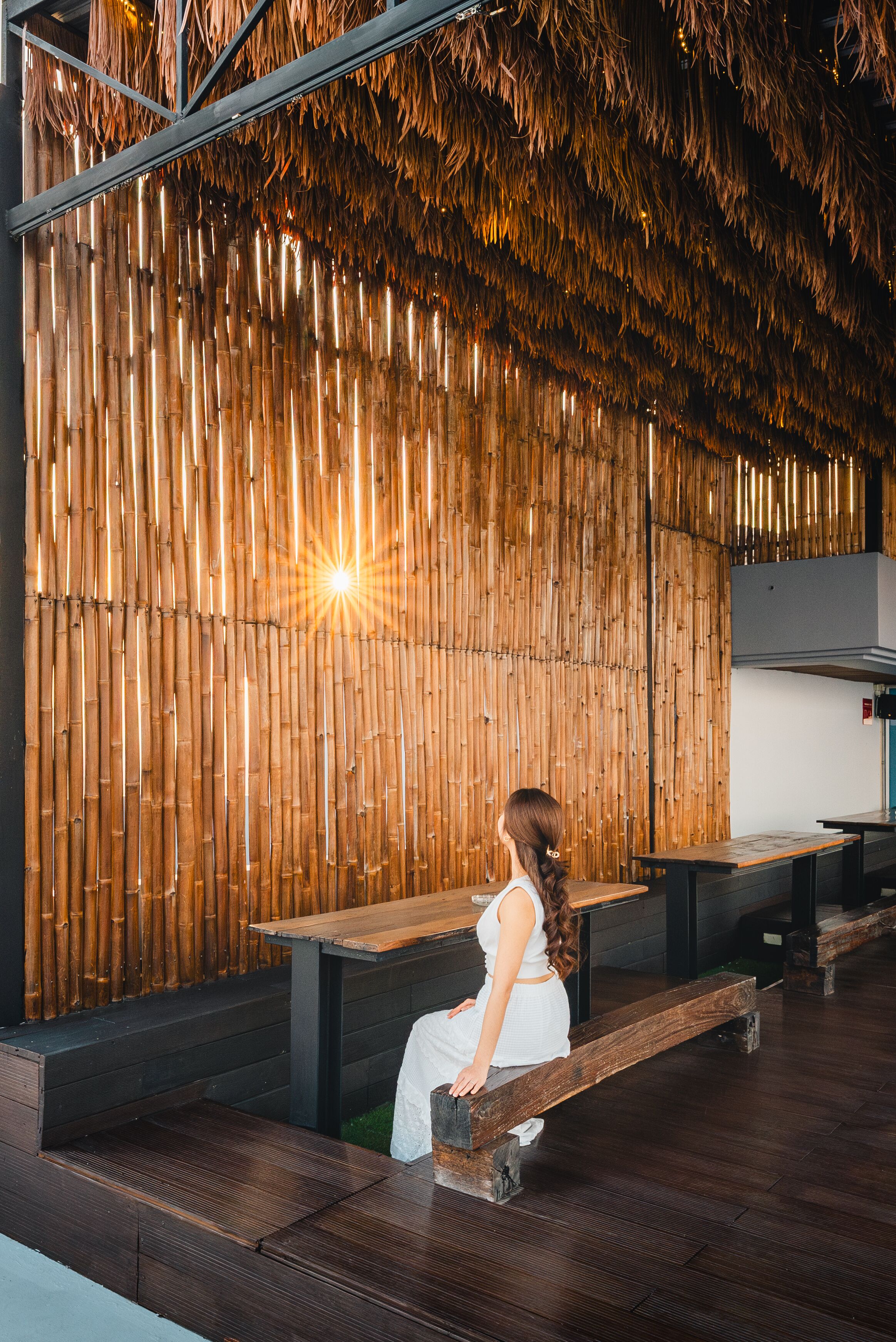 A woman sitting on a wooden bench in a modern space with a bamboo wall and a bright light source.