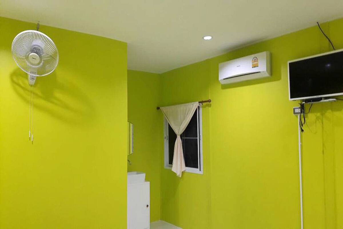 Una habitación con una pared verde brillante, un ventilador blanco, un equipo de aire acondicionado blanco, un lavamanos blanco y una ventana con cortina.