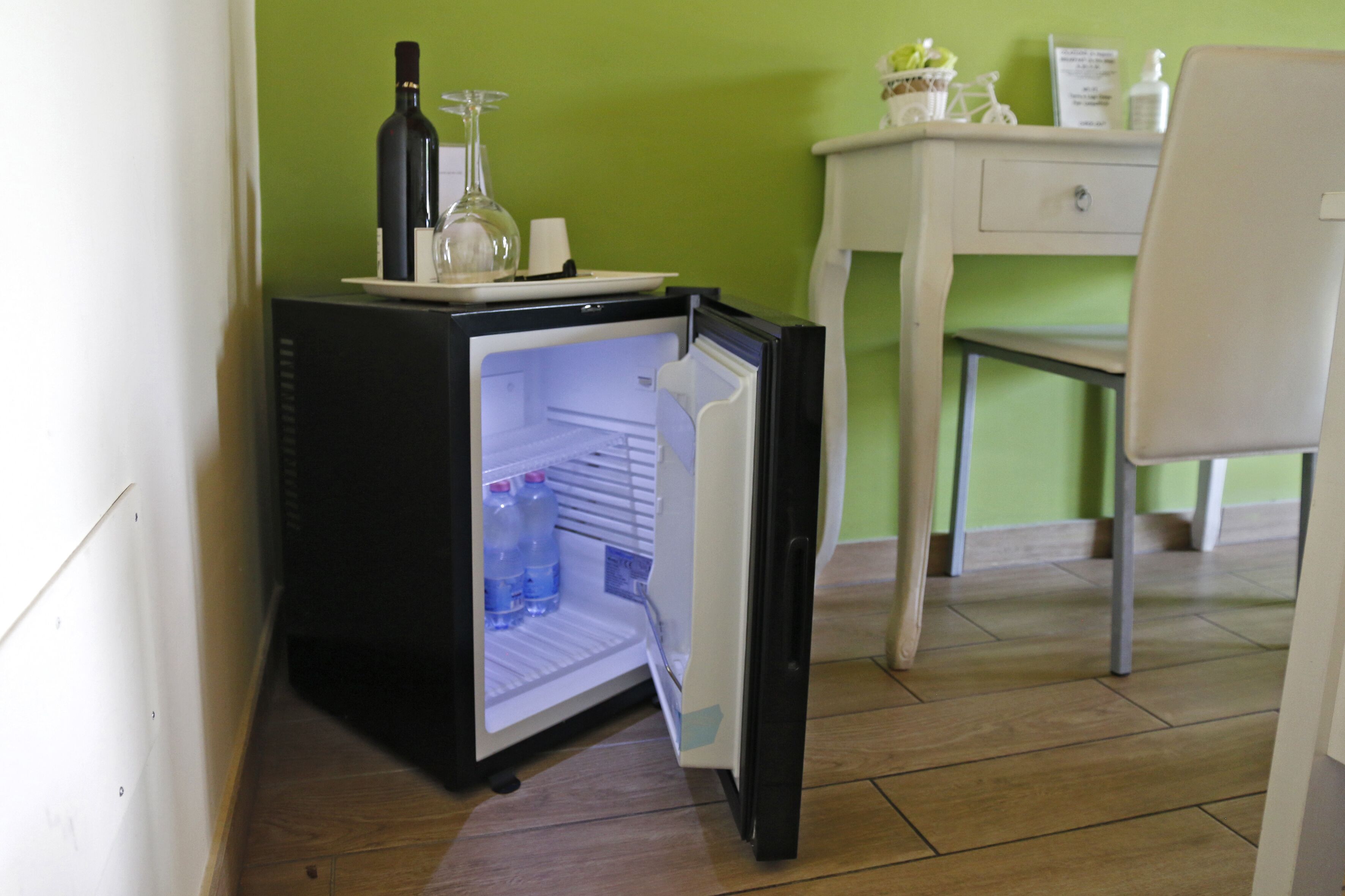 A compact refrigerator with a wine bottle and a glass on top, placed in a room with a green wall and a white chair.