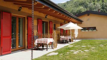 Un edificio con persiane rosse, tavolini all'aperto con ombrelloni bianchi e un'area verde con pietre di passaggio.