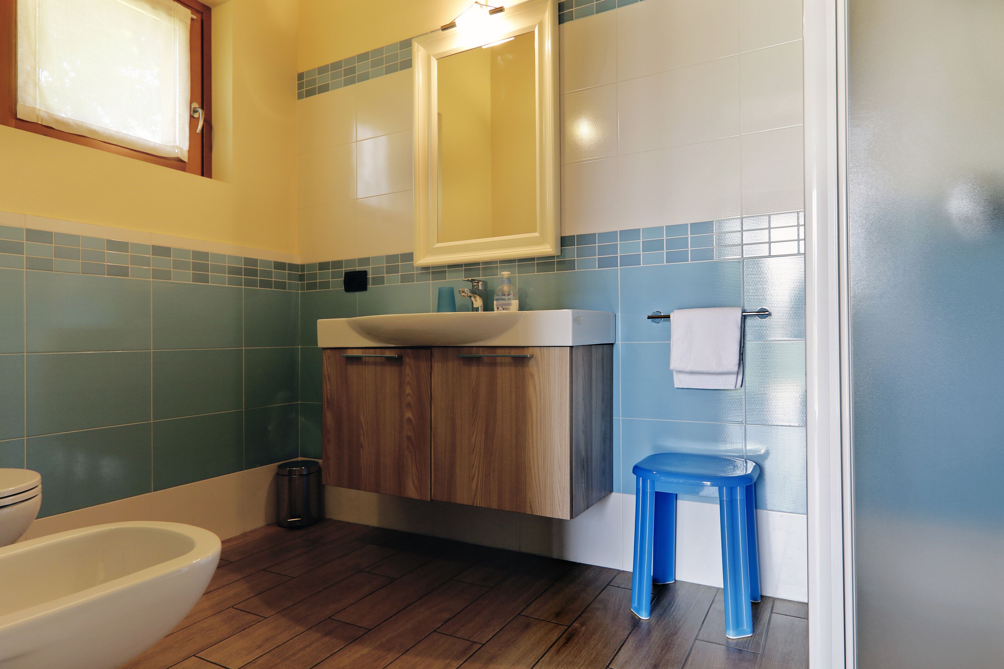 A bathroom with a wooden vanity, a white sink, a blue stool, and a toilet.