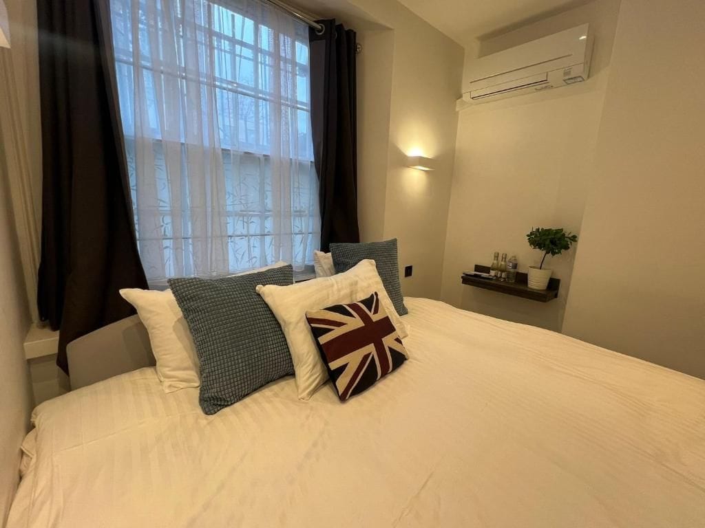 A bedroom with a bed, pillows, a nightstand, a window with curtains, and an air conditioning unit.