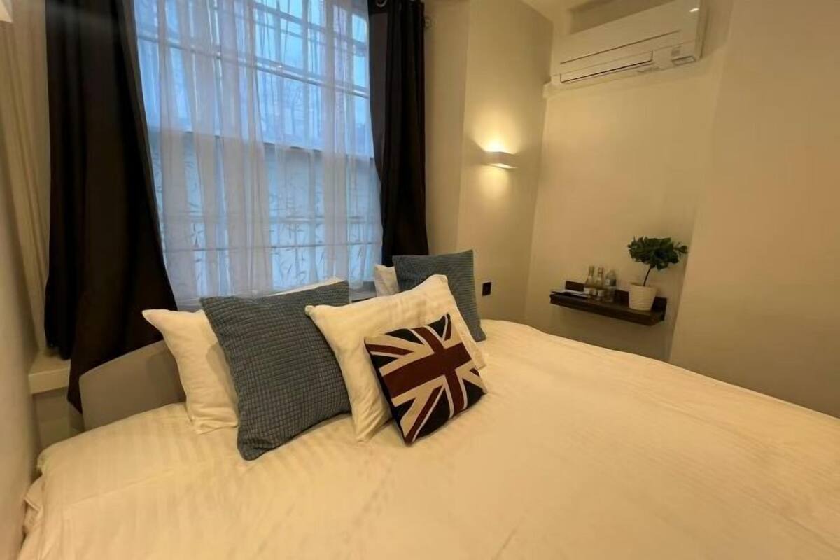 A hotel room with a bed, a window with curtains, a wall-mounted air conditioner, and a small shelf with toiletries.