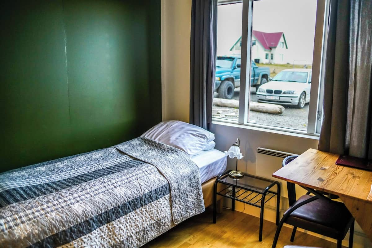 Un lit simple avec une couverture à motifs, une petite table avec une lampe, une chaise et une fenêtre donnant sur la rue, avec vue sur les véhicules et un immeuble.
