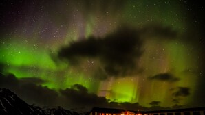 Un bâtiment à l’extérieur sombre, dont les fenêtres sont illuminées, se dressant sur fond de ciel étoilé et d’aurore boréale verdoyante.