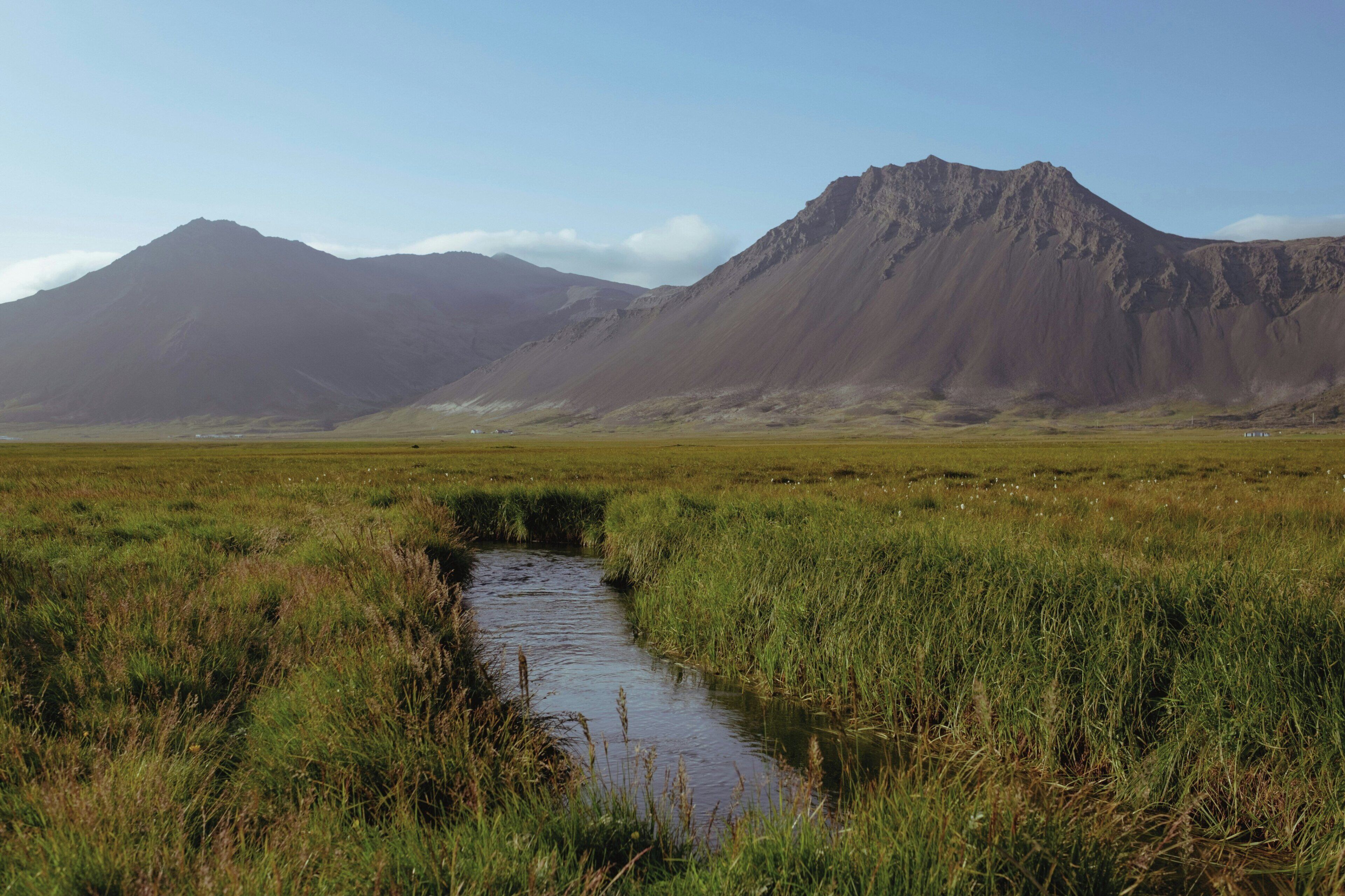 Un paisaje natural con un arroyo, un campo cubierto de hierba y montañas.