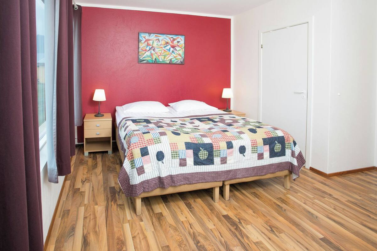 Une chambre avec un plancher en bois, un lit recouvert d’une courtepointe, deux tables de chevet avec des lampes, un mur rouge et une porte.