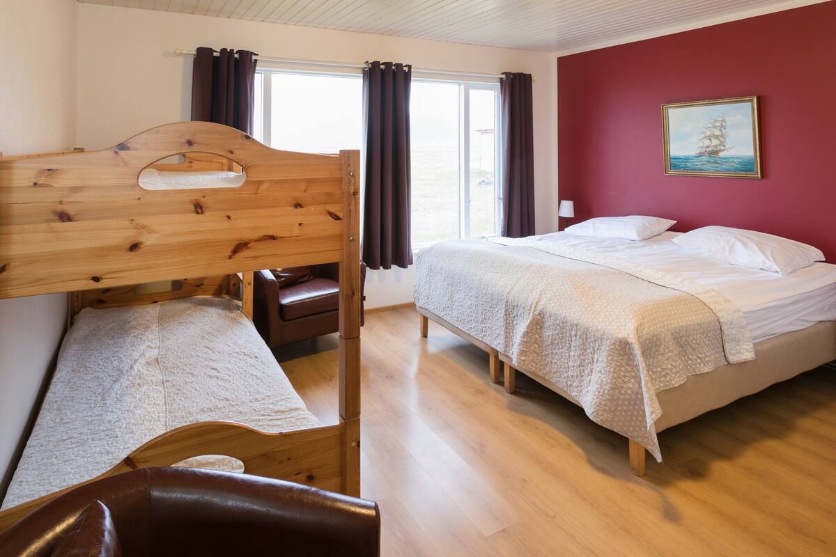 Une chambre avec un lit superposé, un lit simple, un plancher en bois, une fenêtre avec des rideaux et un tableau au mur.