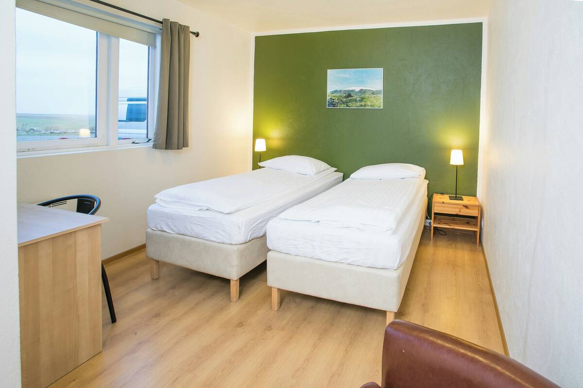 Une chambre avec deux lits, un bureau, une chaise, une fenêtre donnant sur un beau paysage et une lampe fixée au mur.