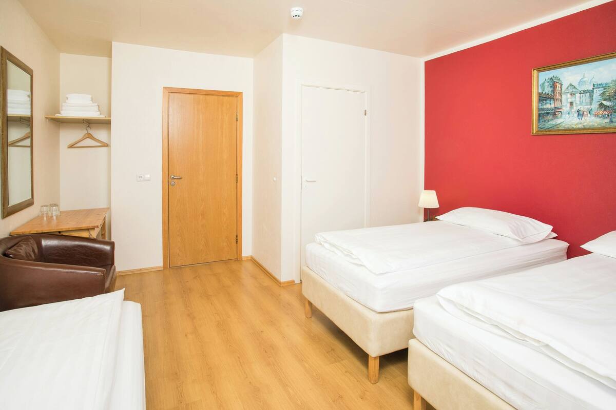 Habitación de hotel con piso de madera, dos camas, un sillón de cuero marrón, una puerta de madera y una pared roja con un cuadro enmarcado.