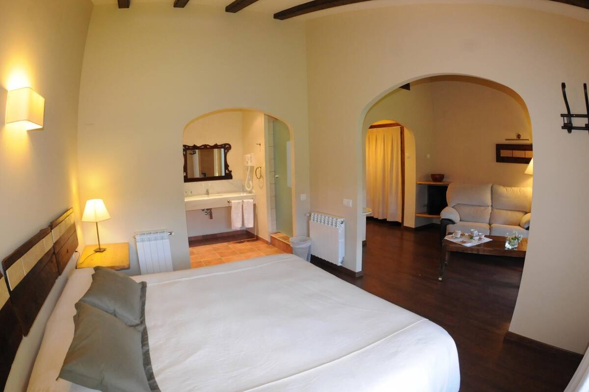 A bedroom with a large bed, a bathroom, and a living area with a sofa and table.