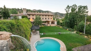 A swimming pool with a stone structure, a well-maintained lawn, and a large building in the background. - Hotel Rural Font del Pas (Beceite)