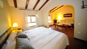 A hotel room with a large bed, a sofa, and a small table. There is a window with curtains and a lamp on a bedside table. - Hotel Rural Font del Pas (Beceite)