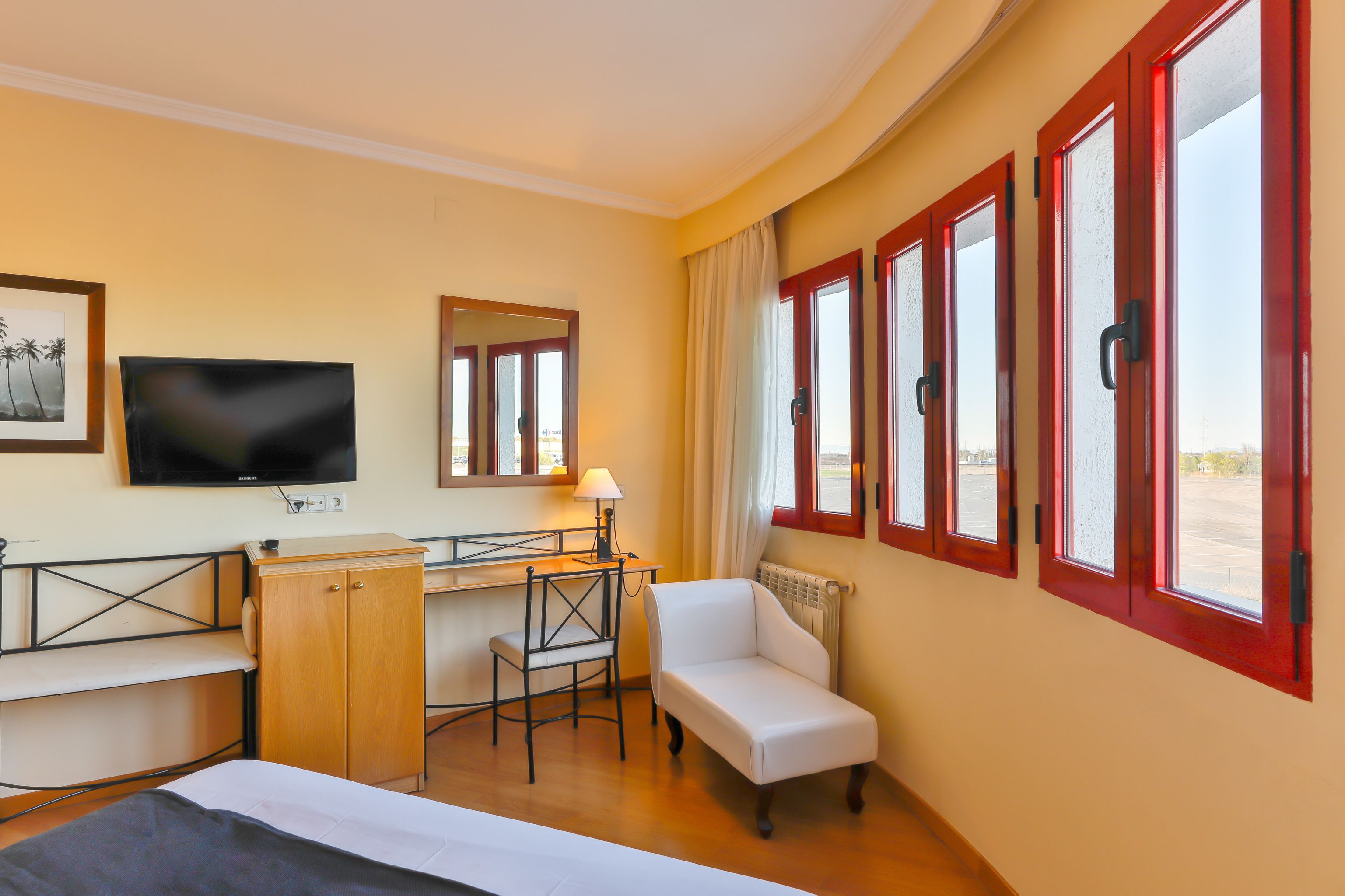 A hotel room with a bed, a desk, a chair, a TV, and a window with red frames.