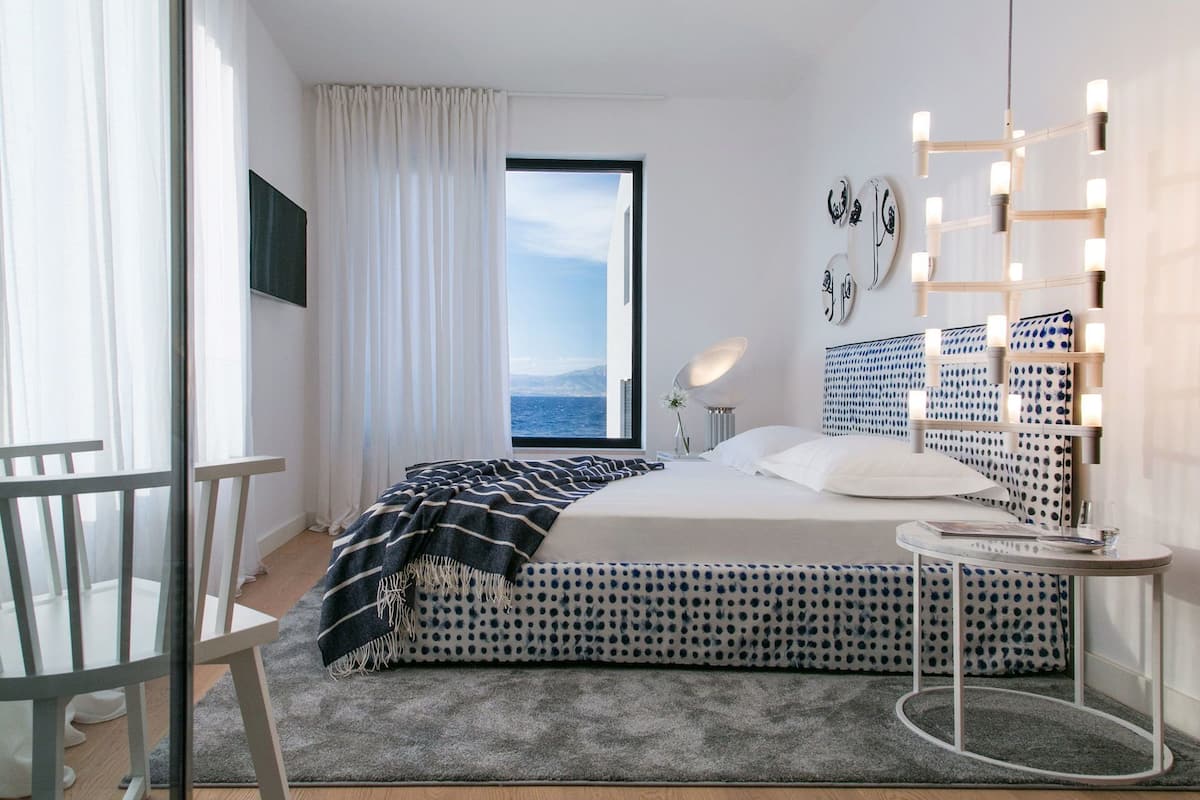 Ein modernes Schlafzimmer mit einem großen Bett, einem Nachttisch, einem Sessel, Meerblick und einem beleuchteten Spiegel.