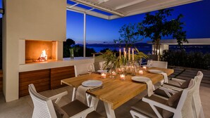 An outdoor dining area with a wooden table, white chairs, candles, and a fire feature. - Omega Villas Complex (Sutivan)