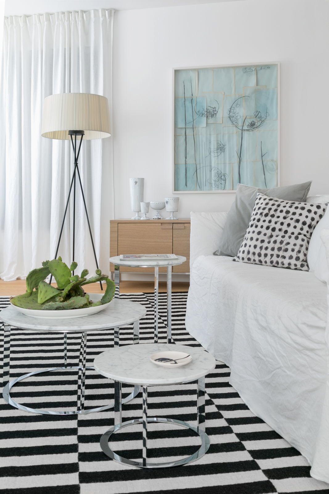 Une chambre d’hôtel moderne avec un lit blanc, un tapis à rayures noires et blanches, une table basse ronde, un lampadaire et un tableau au mur.