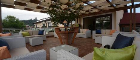 Un patio al aire libre con mobiliario de mimbre blanco, cojines verdes y azules, y una mesa de madera central con una planta en maceta.