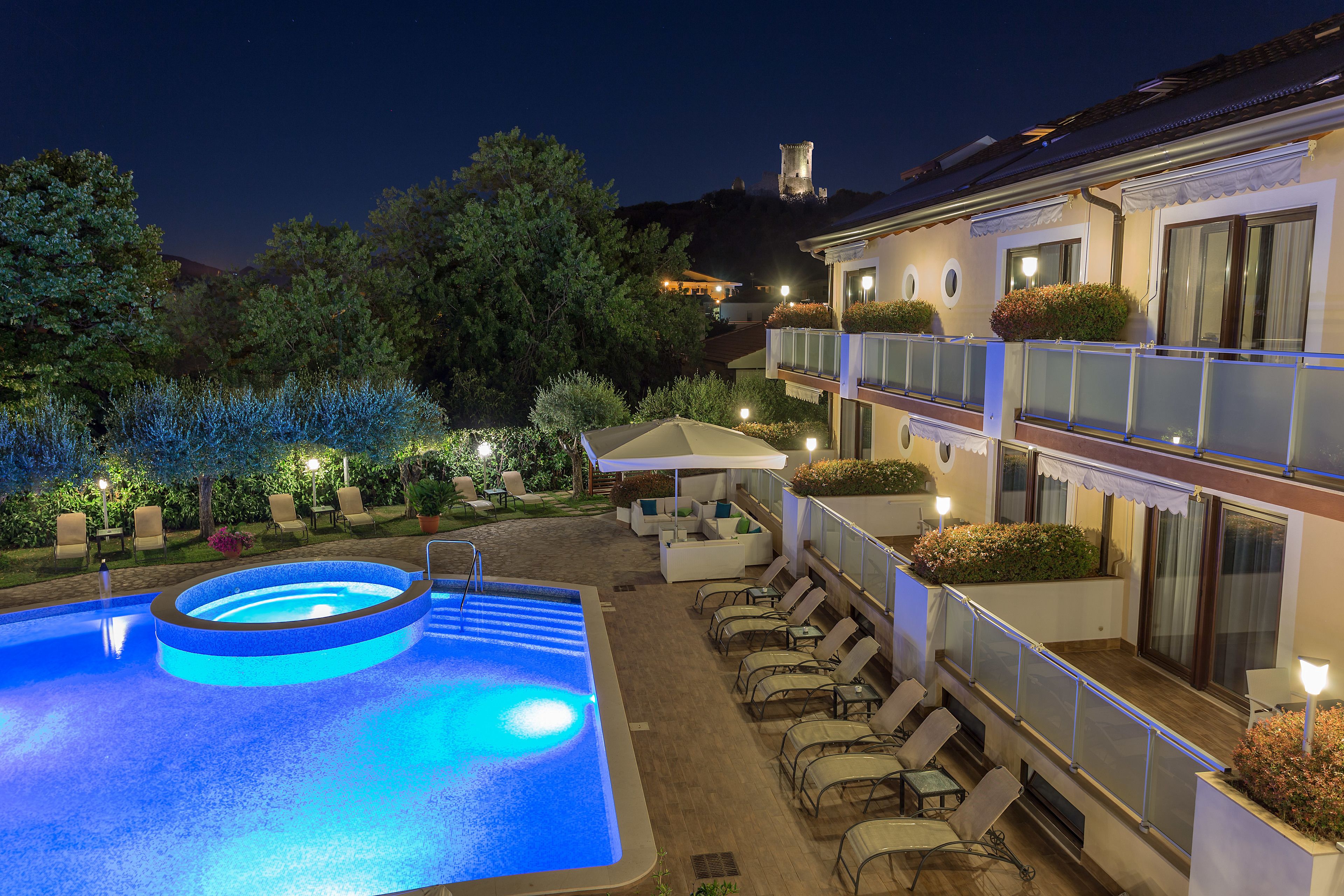 Piscina con sillones de descanso y una zona de estar cubierta, rodeada de vegetación y un edificio con balcón.