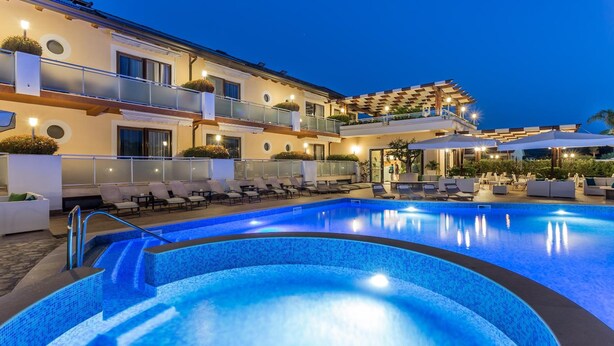 A swimming pool area with lounge chairs and umbrellas, surrounded by a building with balconies and outdoor seating. - Hotel Porta Rosa (Ascea)