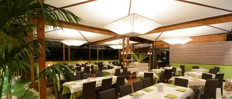 An indoor dining area with multiple tables and chairs, a ceiling fan, and a green artificial plant.
