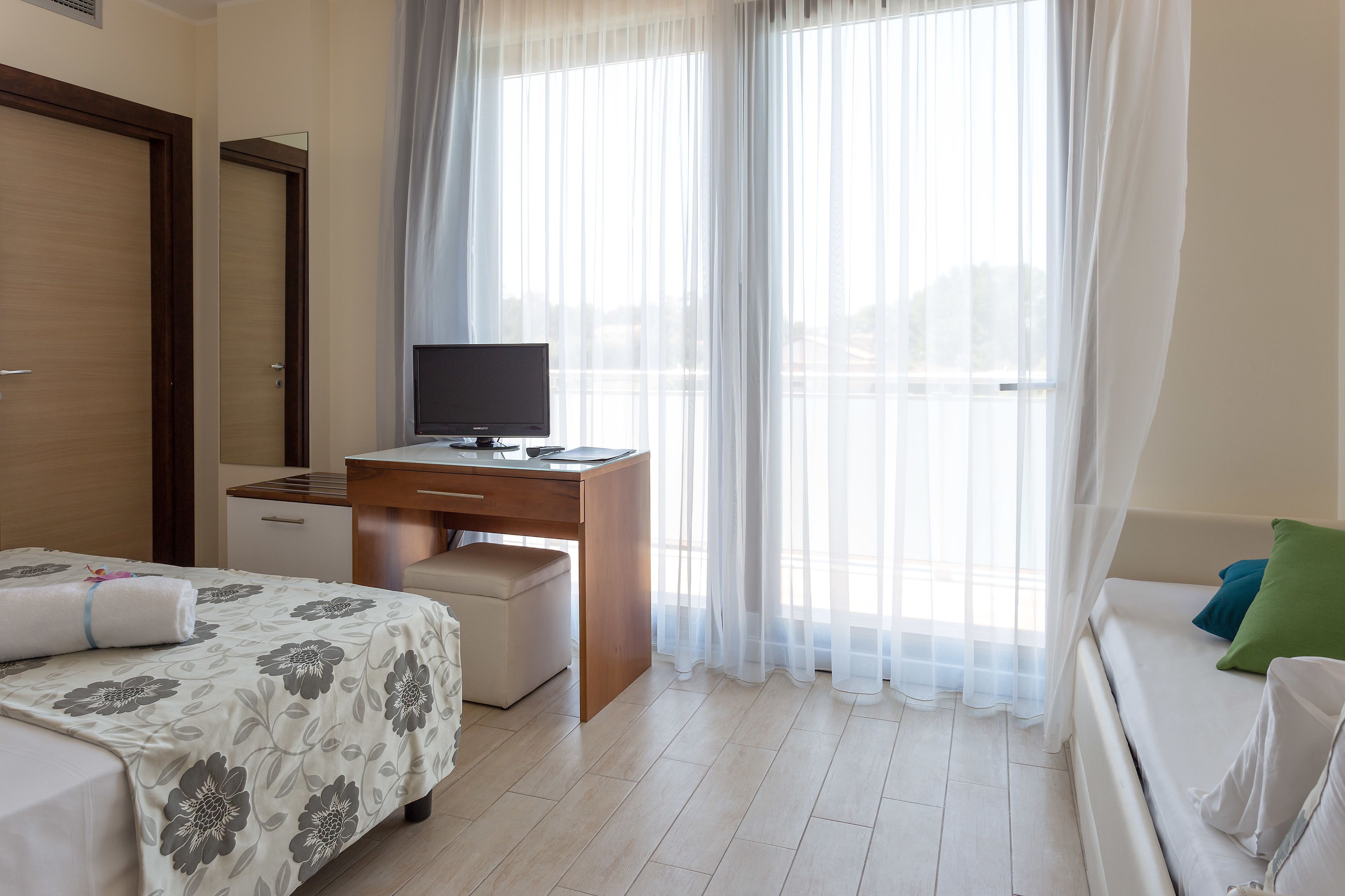 Habitación de hotel con cama, escritorio con computadora, ventana con cortinas translúcidas y sofá con almohadones.