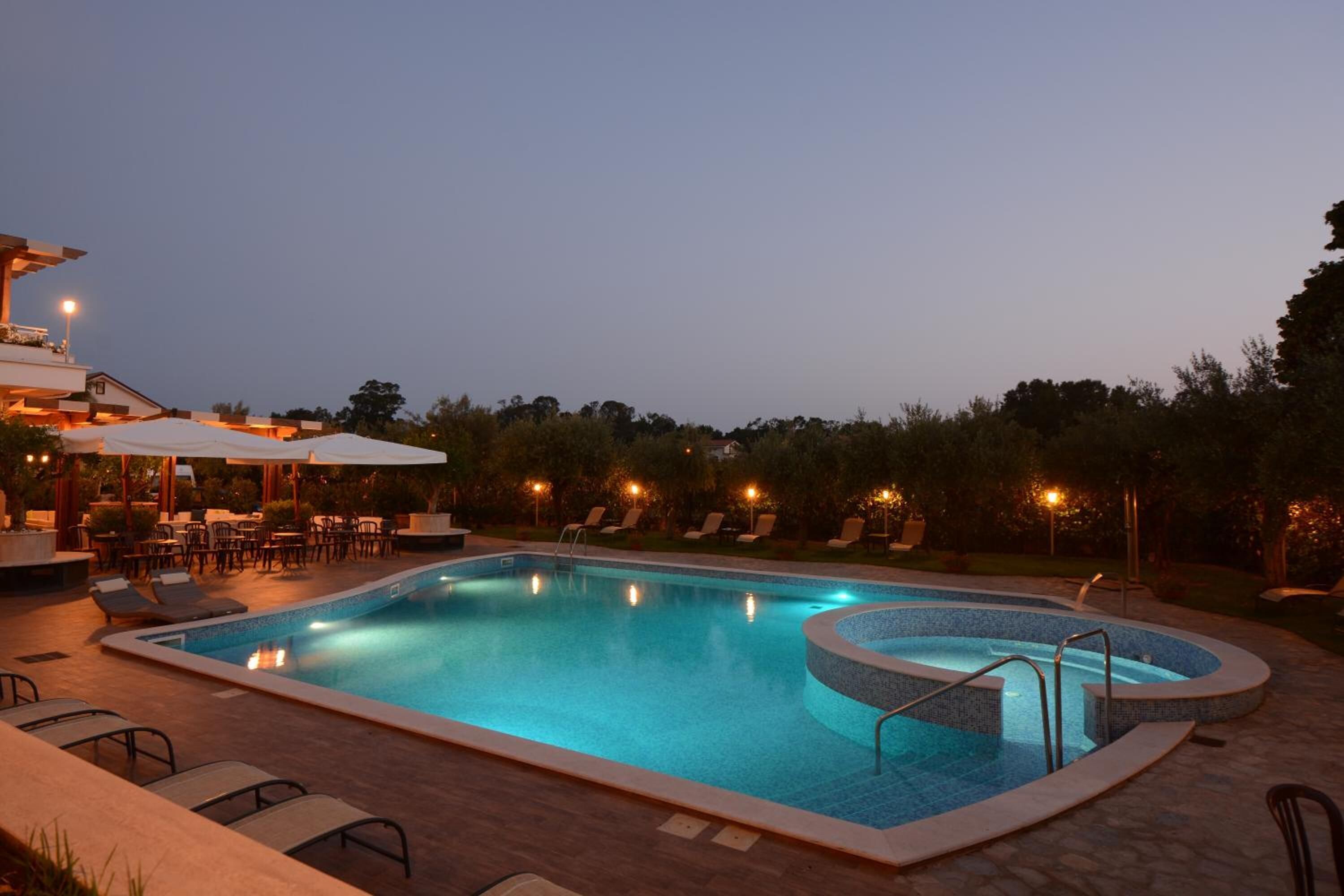 A rectangular outdoor swimming pool with a circular hot tub, surrounded by lounge chairs and tables, and a covered seating area with umbrellas.