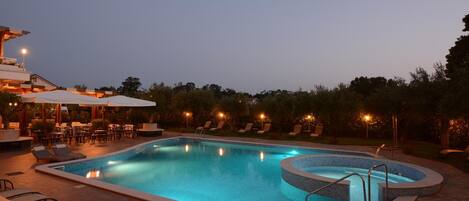 A rectangular outdoor swimming pool with a circular hot tub, surrounded by lounge chairs and tables, and a covered seating area with umbrellas.
