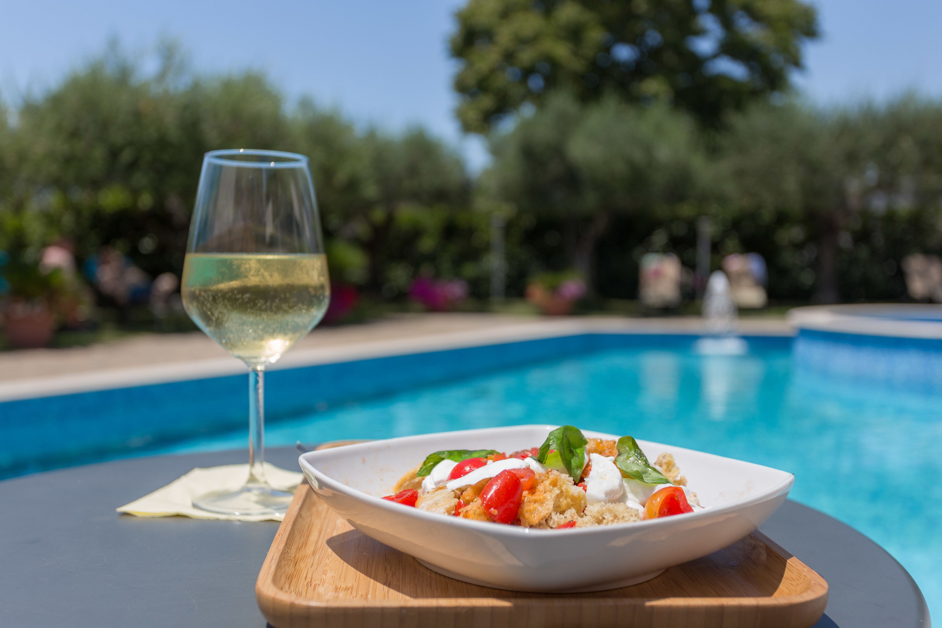 Mesa de jantar com um prato de comida e um copo de vinho branco à beira da piscina.