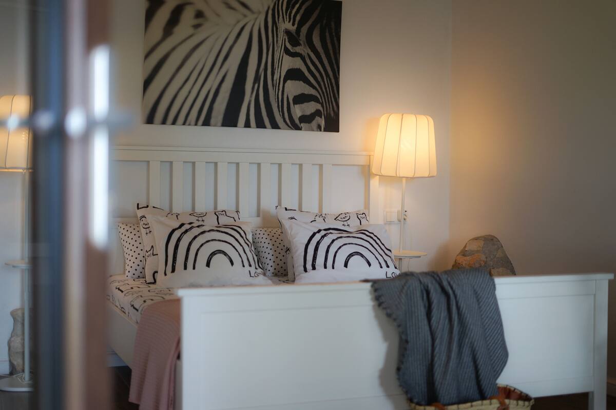 Ein Schlafzimmer mit einem weißen Kopfteil, gemusterten Kissen, Wandkunst mit Zebra-Muster und zwei Nachttischlampen.