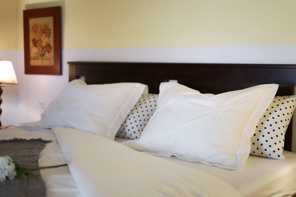 Ein ordentlich bezogenes Bett mit weißen und gepunkteten Kissen, einem Holz-Kopfteil und einem gerahmten Blumenbild an der Wand.