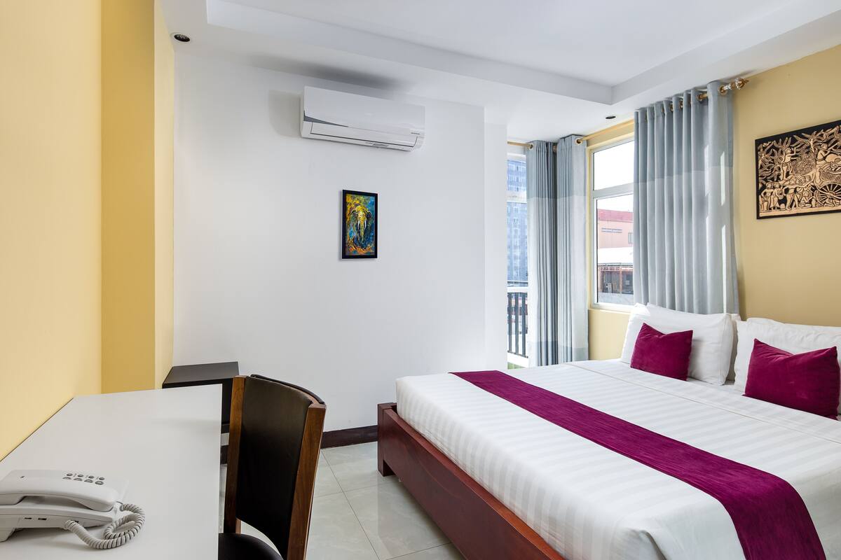 A hotel room with a bed, a desk with a phone, an air conditioner, and a window with curtains.