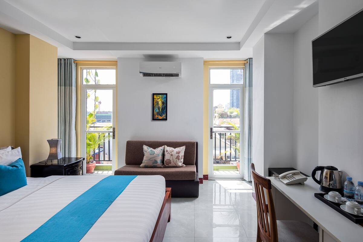 A hotel room with a bed, a sofa, a desk with a phone and a kettle, and a TV on the wall.