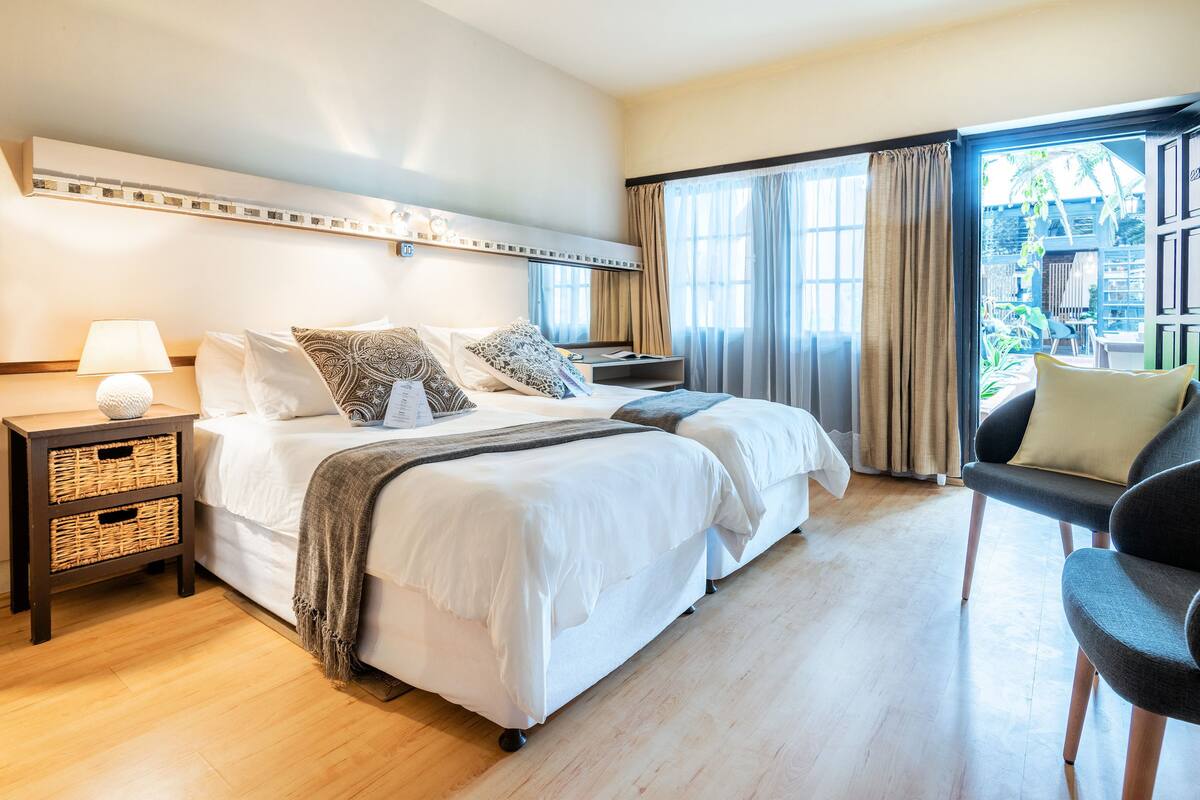 A hotel room with two beds, a bedside table with a lamp, a chair, and a view of the outdoors.