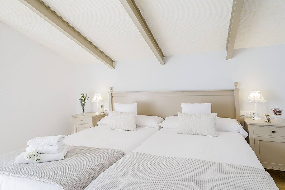 Quarto de hotel com duas camas, uma mesa de cabeceira com abajur e uma mesinha de apoio com um vaso de flores.