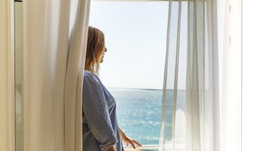 A person standing by a window with sheer curtains, overlooking a view of the sea. - Hotel Gastronómico La Breña (Barbate)