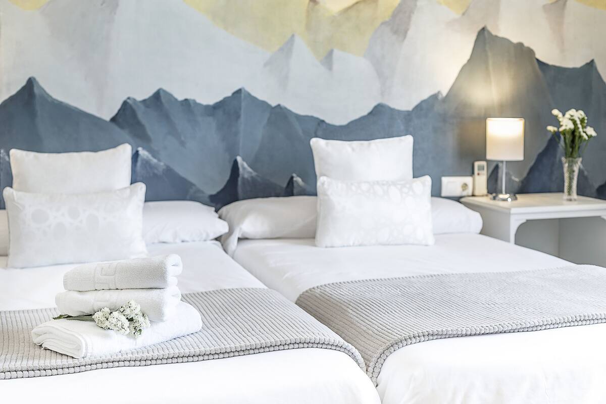 Quarto de hotel com duas camas, roupa de cama branca, painel de parede com tema de montanhas, abajur e um vaso com flores.