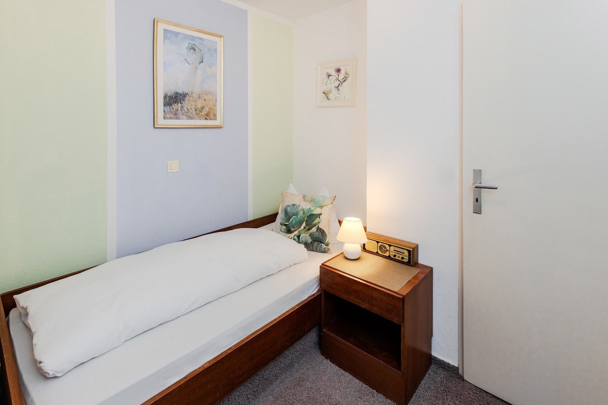 Un petit lit bien fait, avec un drap et un oreiller blancs, une table de chevet avec une lampe, et une porte sur la droite.
