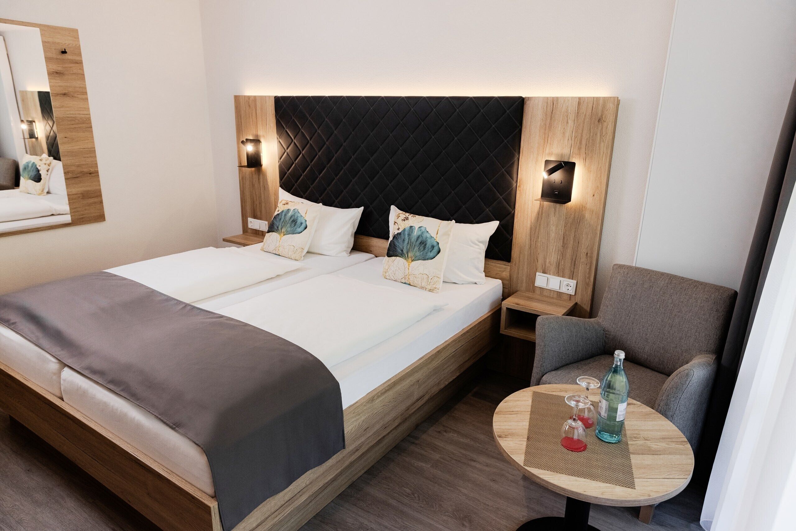 A modern hotel room with a large bed, a wooden headboard, bedside tables, a seating area with a small round table, and a mirror on the wall.