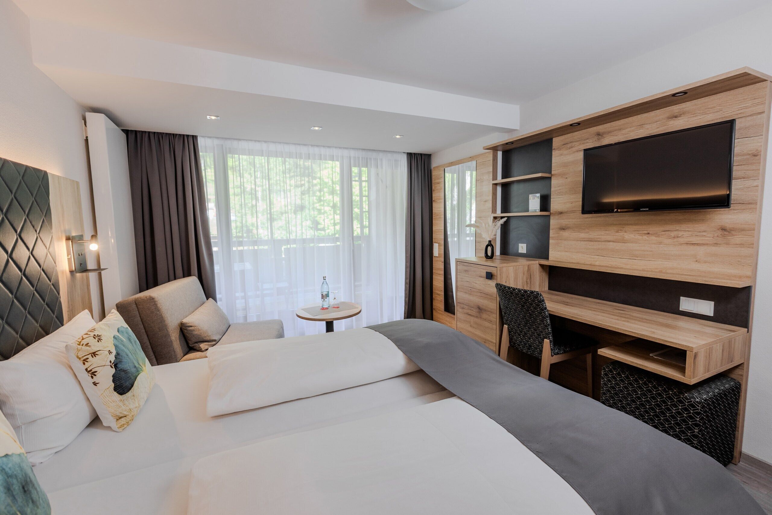 Ein modernes Hotelzimmer mit einem großen Bett, einem an einer Holzwand montierten Fernseher, einem kleinen runden Tisch und einer Sitzecke mit einem Sessel.